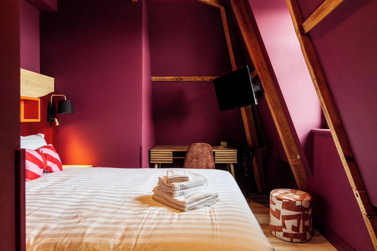 A bedroom with a bed, a wooden desk, a chair, a television, and a small ottoman.
