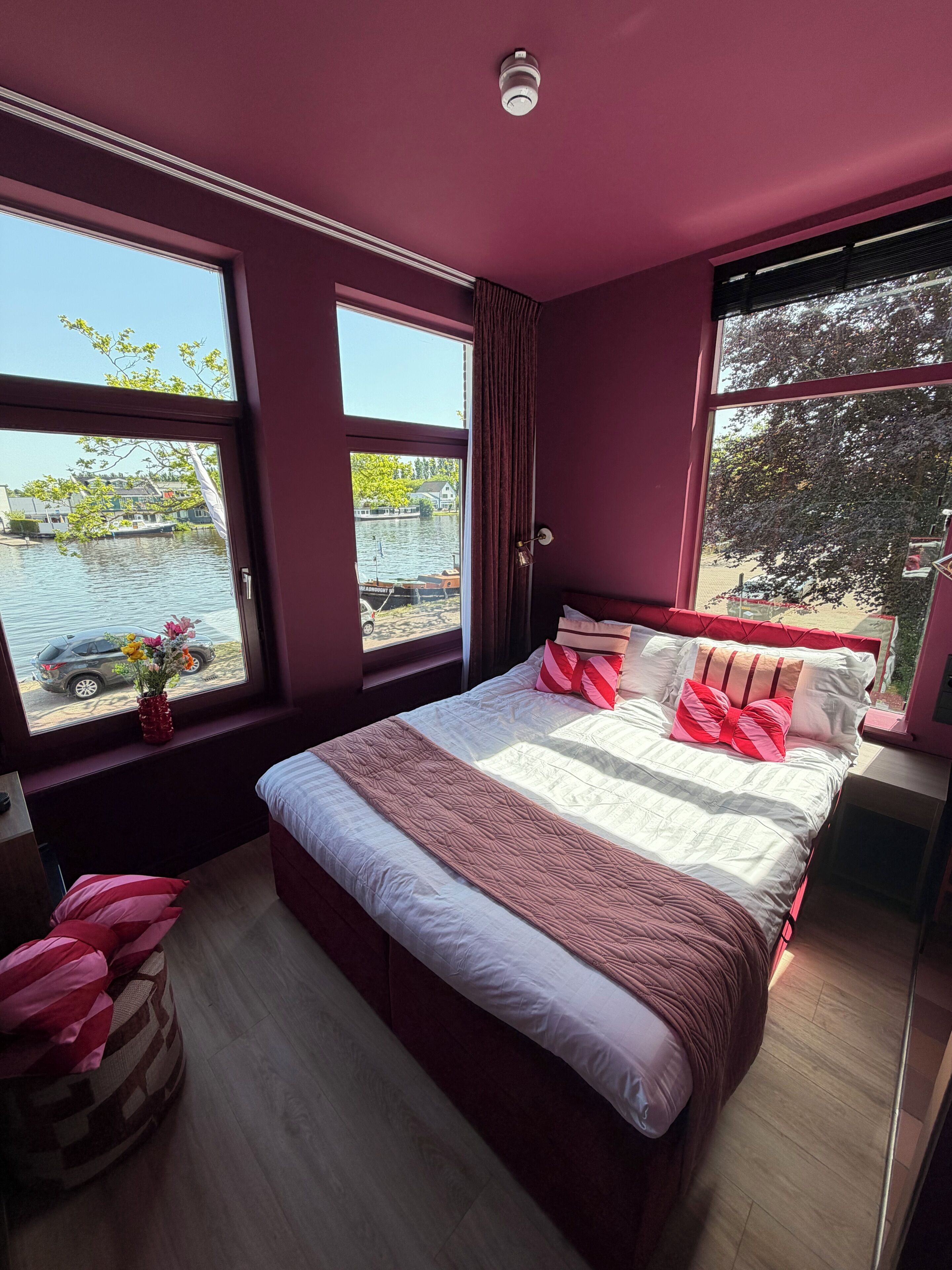 A bedroom with a large bed, a window with a view of water and boats, and a red vase with flowers on the windowsill.