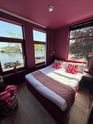 Une chambre avec un grand lit, une fenêtre donnant sur l’eau et les bateaux, et un vase rouge orné de fleurs, posé sur le rebord de la fenêtre.