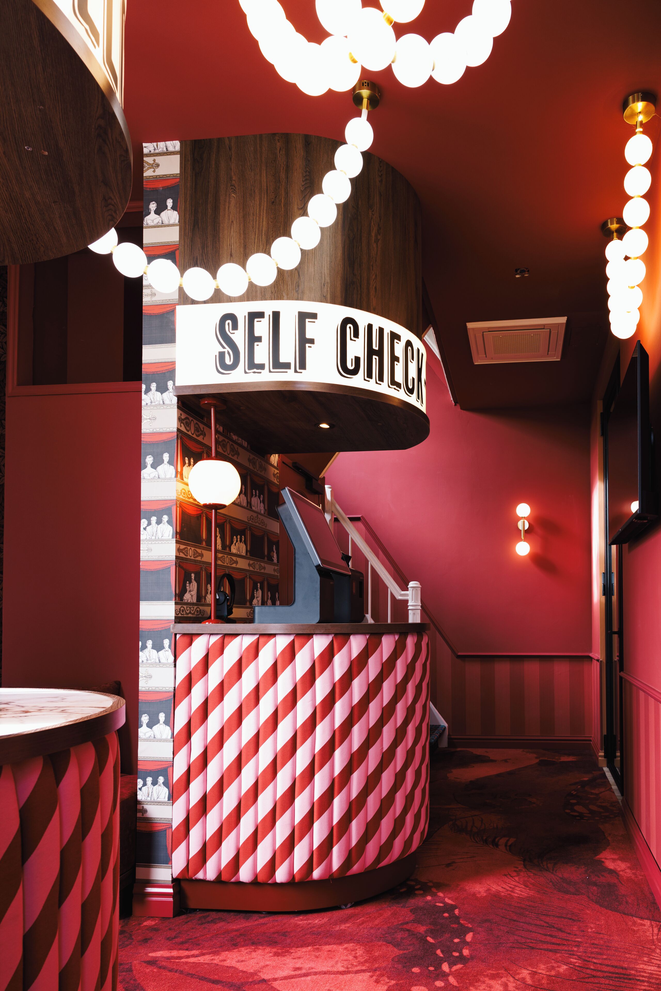A self-checkout station with a red and white striped counter, a sign above it, and a staircase leading to an upper level.