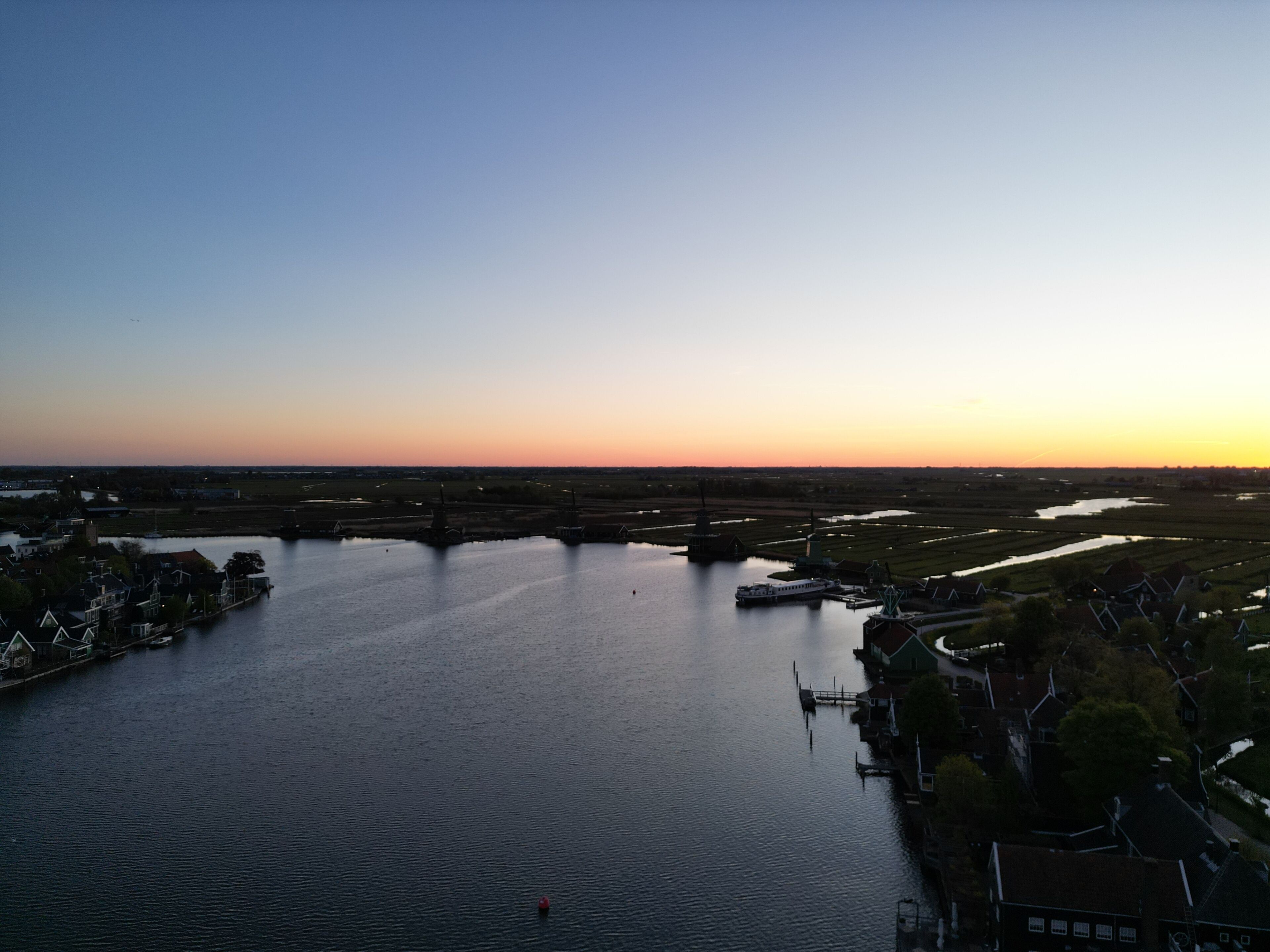 Een rivier met een brug, huizen langs de oever en uitgestrekte velden in de verte.