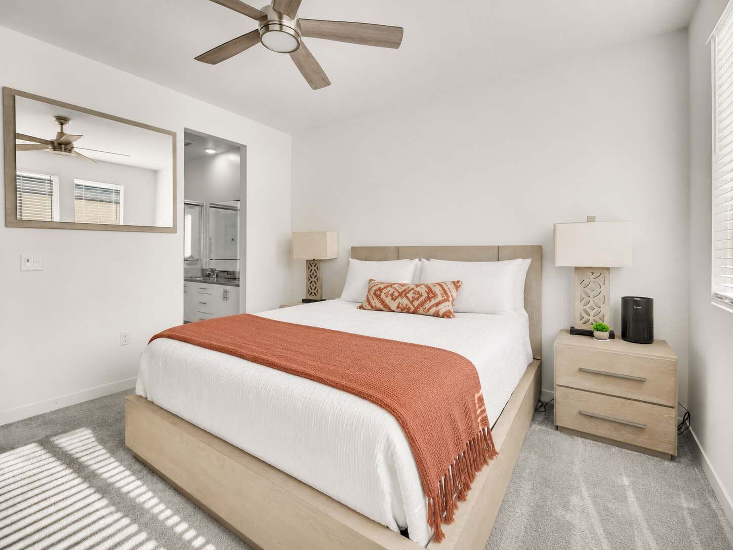 A hotel room with a bed, a ceiling fan, a nightstand, a lamp, a mirror, and a window with blinds.