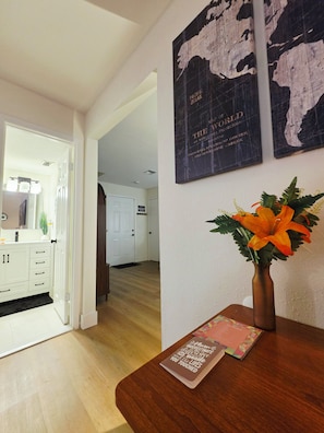 A hallway with a white door leading to a bathroom, a wooden table with a vase of flowers, and two framed maps on the wall. - Welcome to our beautiful centrally located home with a private conversation view (Lakeland)