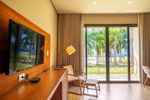 A modern hotel room with a large flat-screen TV, a wooden desk, a comfortable chair, a small table, and a view of palm trees and a golf course through large windows.