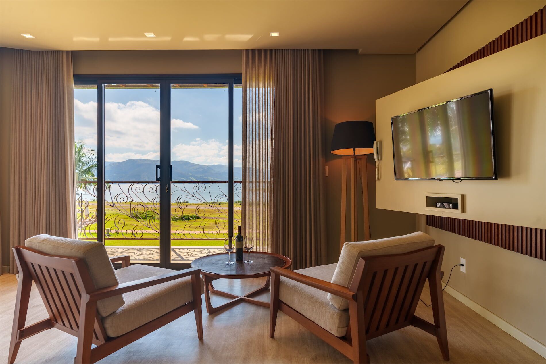 A hotel room with a balcony, two armchairs, a round table, a TV mounted on the wall, and a lamp.
