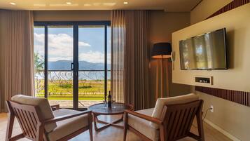 A hotel room with a balcony, two armchairs, a round table, a TV mounted on the wall, and a lamp.