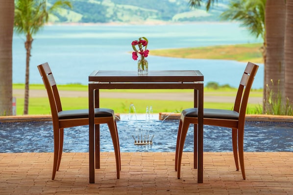 A table with two chairs and a vase of flowers, overlooking a body of water with a fountain in the center.