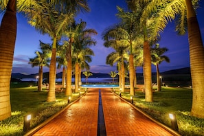 A paved pathway lined with palm trees leading to a pool area, with a view of a body of water and mountains in the distance.
