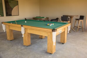 Una mesa de billar con superficie de fieltro verde, rodeada de sillas y una mesita.