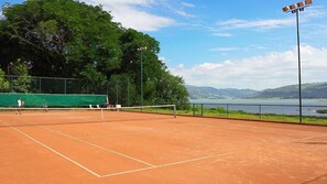 Una cancha de tenis al aire libre con una red, rodeada por una valla, y una vista de un lago y colinas al fondo.