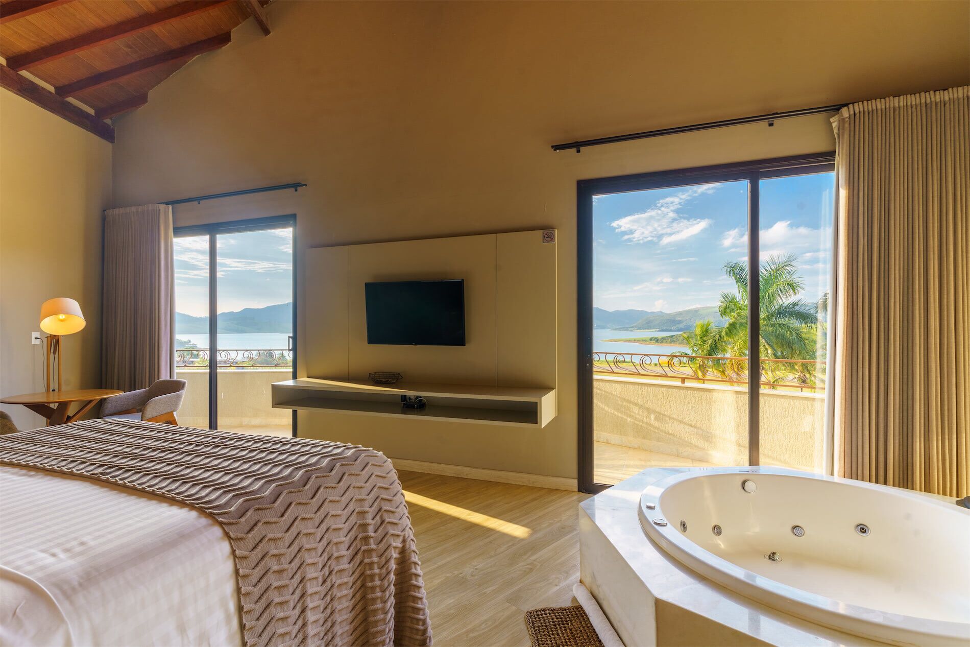 A hotel room with a bed, a TV mounted on the wall, a balcony with a view of the sea, and a bathtub.