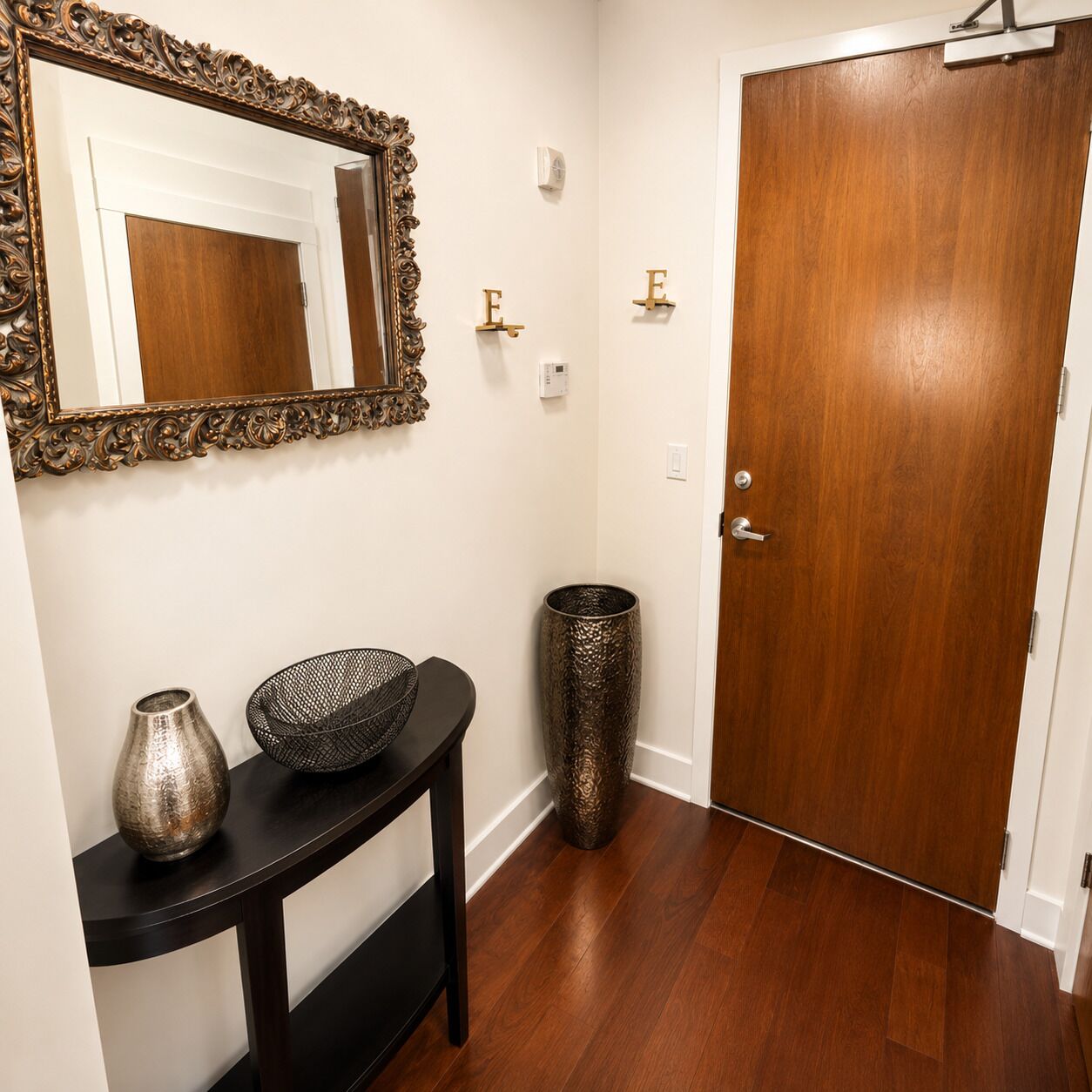 A hallway with a wooden door, a mirror with an ornate frame, a console table with decorative items, and a textured vase.