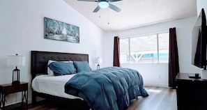 Une chambre à coucher avec un lit, un ventilateur de plafond, une fenêtre avec des rideaux, un tableau au mur et un téléviseur posé sur un meuble.