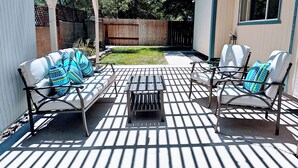 Um pátio com mesa de metal e dois sofás com almofadas estampadas, cerca de madeira ao fundo, e uma casa com janela.