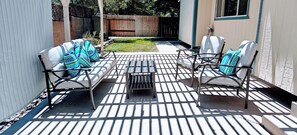 Eine Terrasse mit einem Metalltisch und zwei Sofas mit gemusterten Kissen, ein Holzzaun im Hintergrund und ein Haus mit einem Fenster.