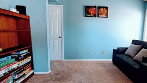 Um quarto com estante, porta, parede azul, sofá preto e dois cartazes com os dizeres 'FUTEBOL' e 'BEISEBOL'.