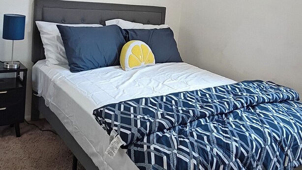 Quarto com cama, mesa de cabeceira, abajur, ventilador de teto e duas imagens penduradas na parede.