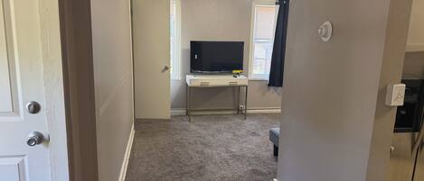 Un couloir recouvert de moquette mène à une chambre dotée d’un ventilateur de plafond, d’un téléviseur posé sur un bureau et d’une fenêtre avec des rideaux.