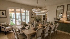 Un espace repas avec une grande table dressée pour six personnes, entourée de chaises, d’un lustre et offrant une vue à travers des portes vitrées.