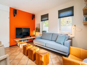 A living room with a TV, sofa, wooden stools, and a window with blinds. - Spa Retreat With Private Sauna (De Koog)