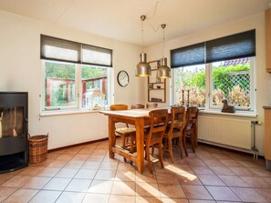 A dining area with a wooden table and chairs, a clock on the wall, and a view of greenery outside. - Spa Retreat With Private Sauna (De Koog)