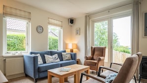 Ett vardagsrum med en blå soffa, ett soffbord i trä, två fåtöljer, en öppen spis och stora fönster med gardiner.