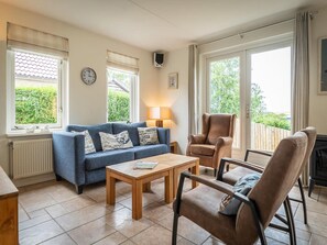A living room with a blue sofa, a wooden coffee table, two chairs, a fireplace, and large windows with curtains. - Spa Retreat With Private Sauna (De Koog)