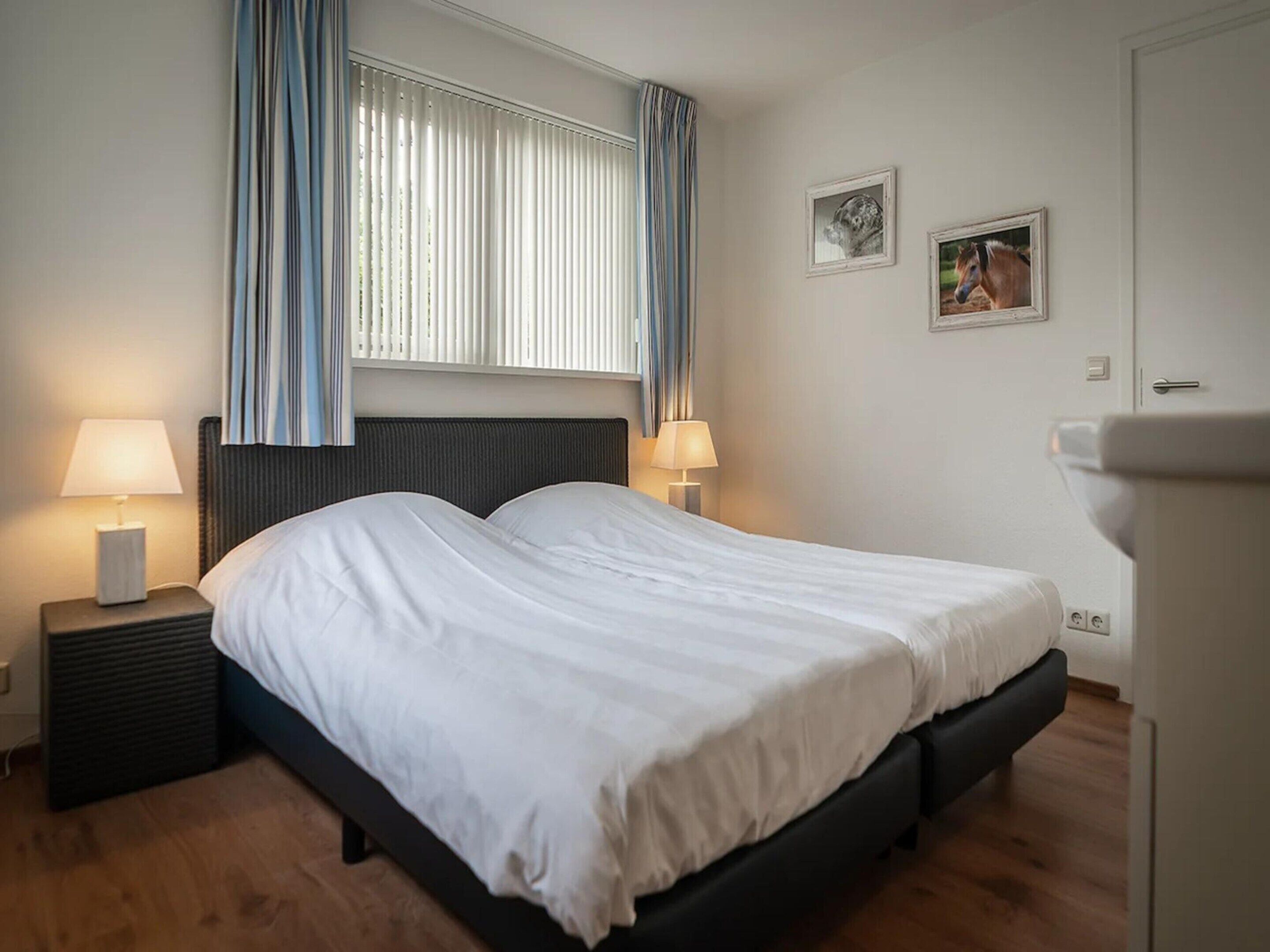 A bedroom with a bed, two bedside tables, a window with curtains, and two wall-mounted pictures.