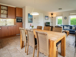 Uma cozinha moderna com armários de madeira, uma mesa de jantar com cadeiras de vime, uma TV e um sofá em uma sala de estar com grandes janelas.