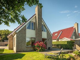 A two-story house with a brick exterior, a red roof, and a small front yard with a flower bed and outdoor furniture.