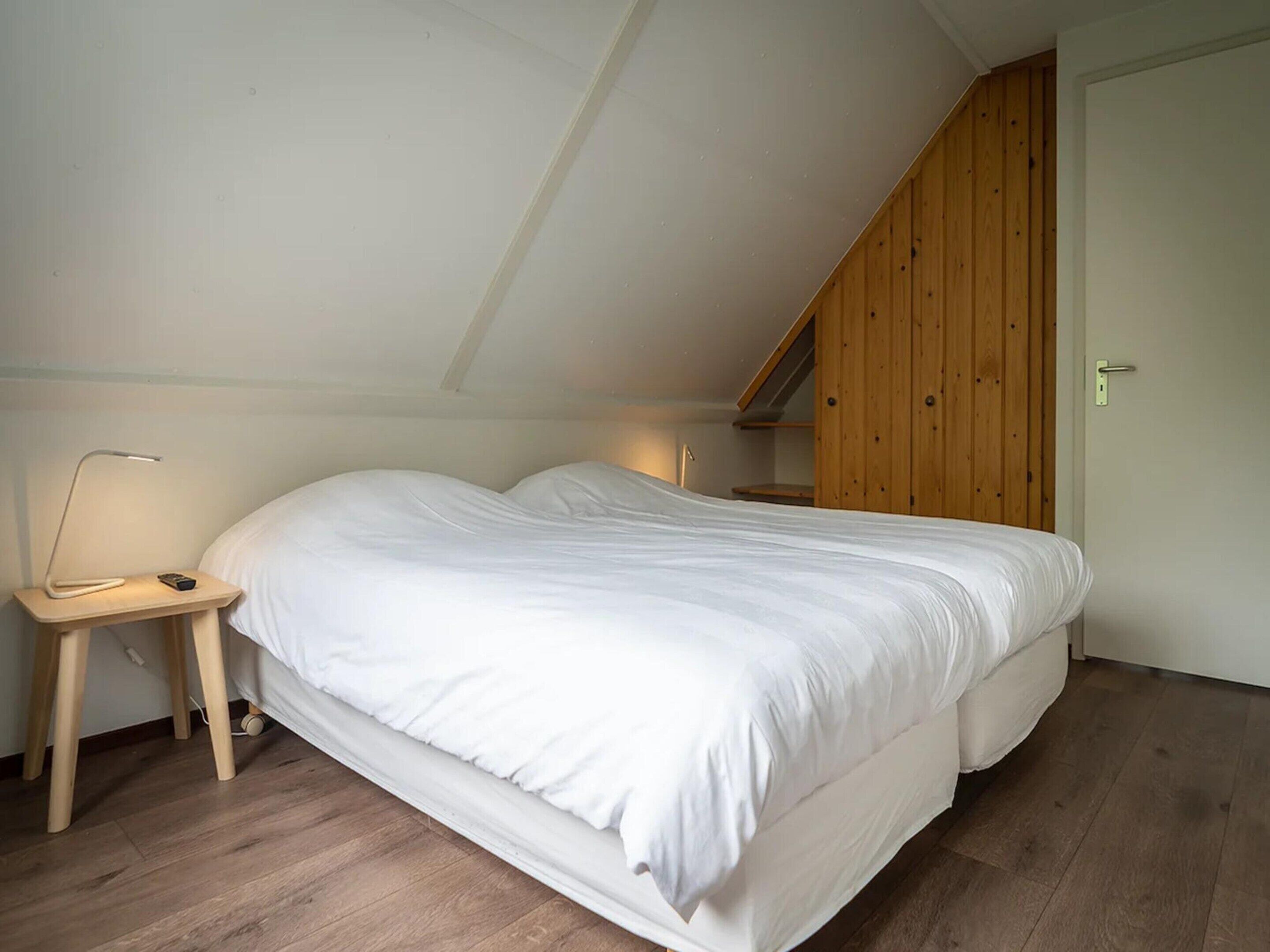 Ein Schlafzimmer mit schrägem Dach, einem Bett, einem Nachttisch mit Lampe und einer Holztür.