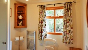 A bathroom with a toilet, a sink, a towel rack, a shelf with a teapot, and a window with floral curtains.