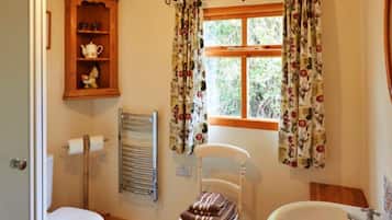 Un baño con inodoro, lavamanos, toallero, estante con tetera y ventana con cortinas florales.