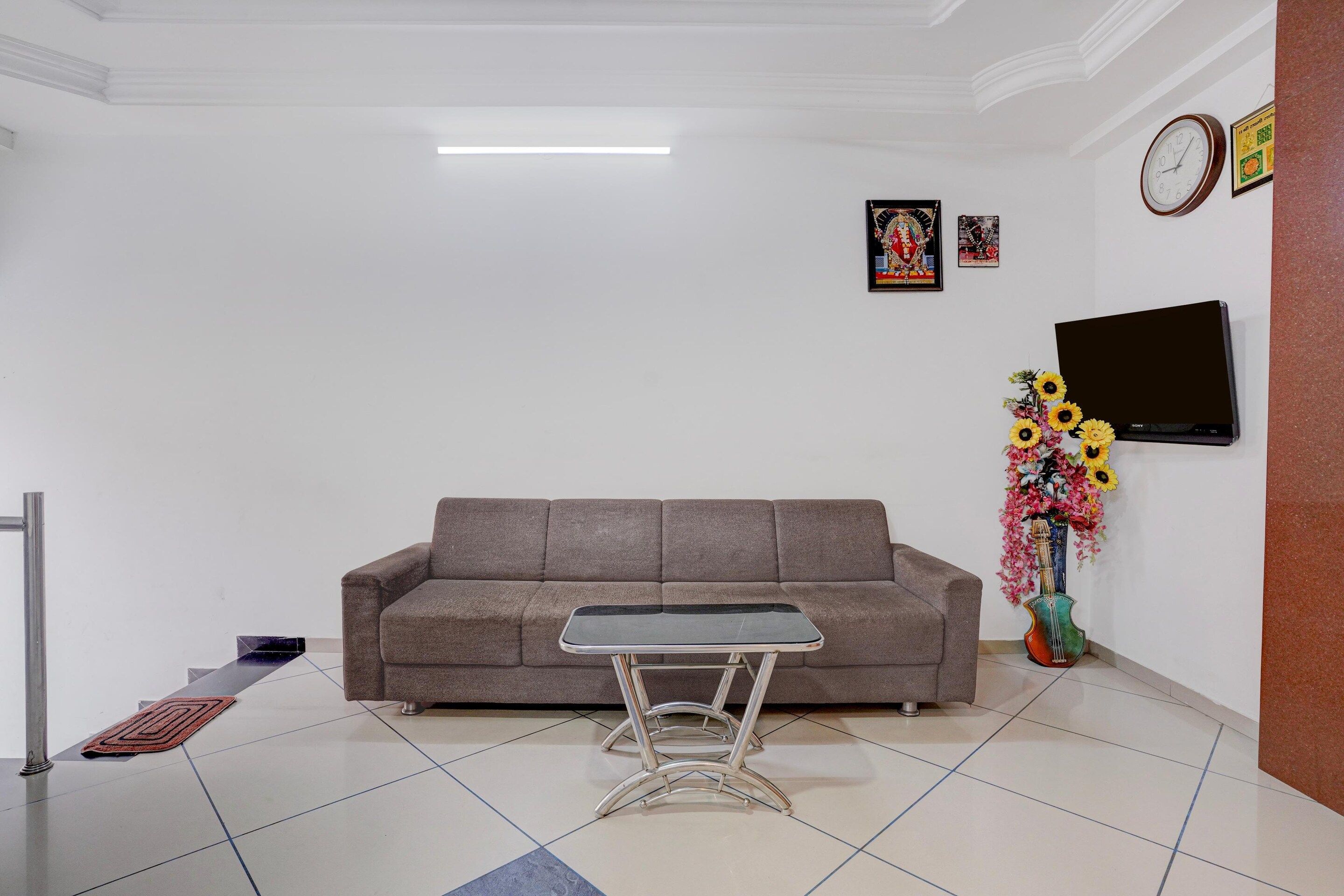 A living room with a sofa, a coffee table, a flat-screen TV, a wall-mounted clock, and a floral arrangement.