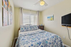 A bedroom with a bed, a ceiling fan, a window with blinds, a bedside table, a TV, and artwork on the walls.