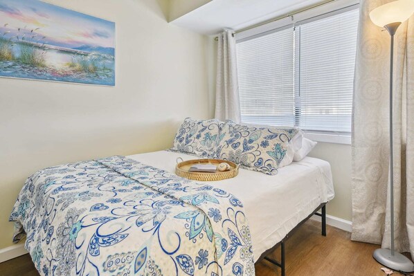 A bedroom with a bed, a bedside table, a lamp, a window with blinds, and a painting on the wall.