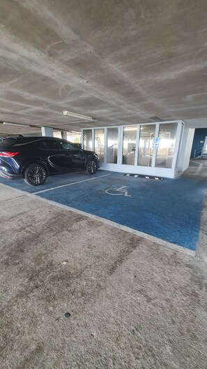 A parking area with a blue handicapped parking space, a black car parked, and a building entrance with glass doors. - Oceanfront Bliss, Top Floor, 2BR, Indoor Pool & Hot Tub, Free Parking, Elevators (North Myrtle Beach)