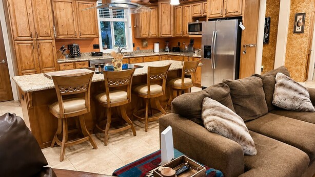 Una cocina con gabinetes de madera, un refrigerador de acero inoxidable, una zona de comedor con taburetes de bar y un sofá marrón con almohadones.