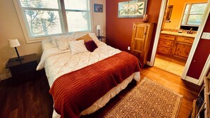 Un dormitorio con cama, mesita de noche, lámpara, ventana, un cuadro, armario y baño visible a través de una puerta abierta.