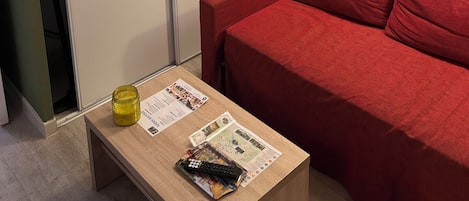 Une pièce avec un canapé rouge, une table basse en bois sur laquelle sont posés des magazines, une lampe et une citation encadrée accrochée au mur.