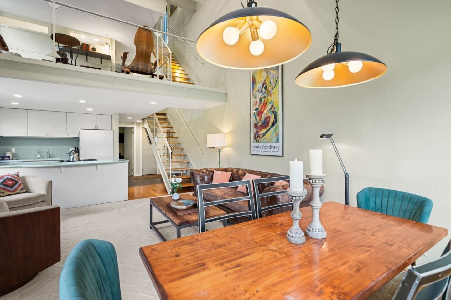 Amplia sala de estar con una mesa de comedor de madera, sillas color turquesa, una escalera y una cocina al fondo.