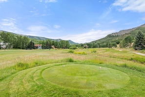 A golf course with a well-maintained fairway, a putting green, and a backdrop of residential buildings and mountains.
