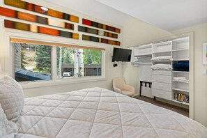 A bedroom with a large bed, a window with a view of trees and a car, a TV mounted on the wall, and a walk-in closet with shelves and hanging space.