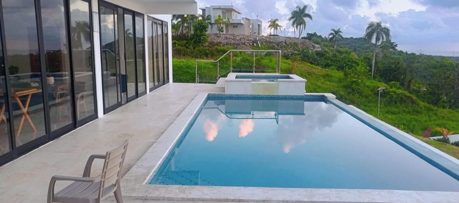 A modern outdoor swimming pool with a glass enclosure, a lounge area with chairs, and a view of a lush green landscape with palm trees.