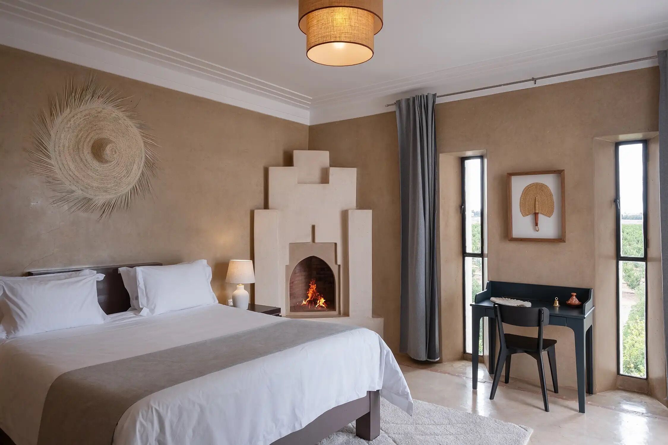 A bedroom with a large bed, a fireplace, a desk with a chair, and a window with curtains.