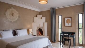 A bedroom with a large bed, a fireplace, a desk with a chair, and a window with curtains.