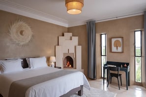 A bedroom with a large bed, a fireplace, a desk with a chair, and a window with curtains.
