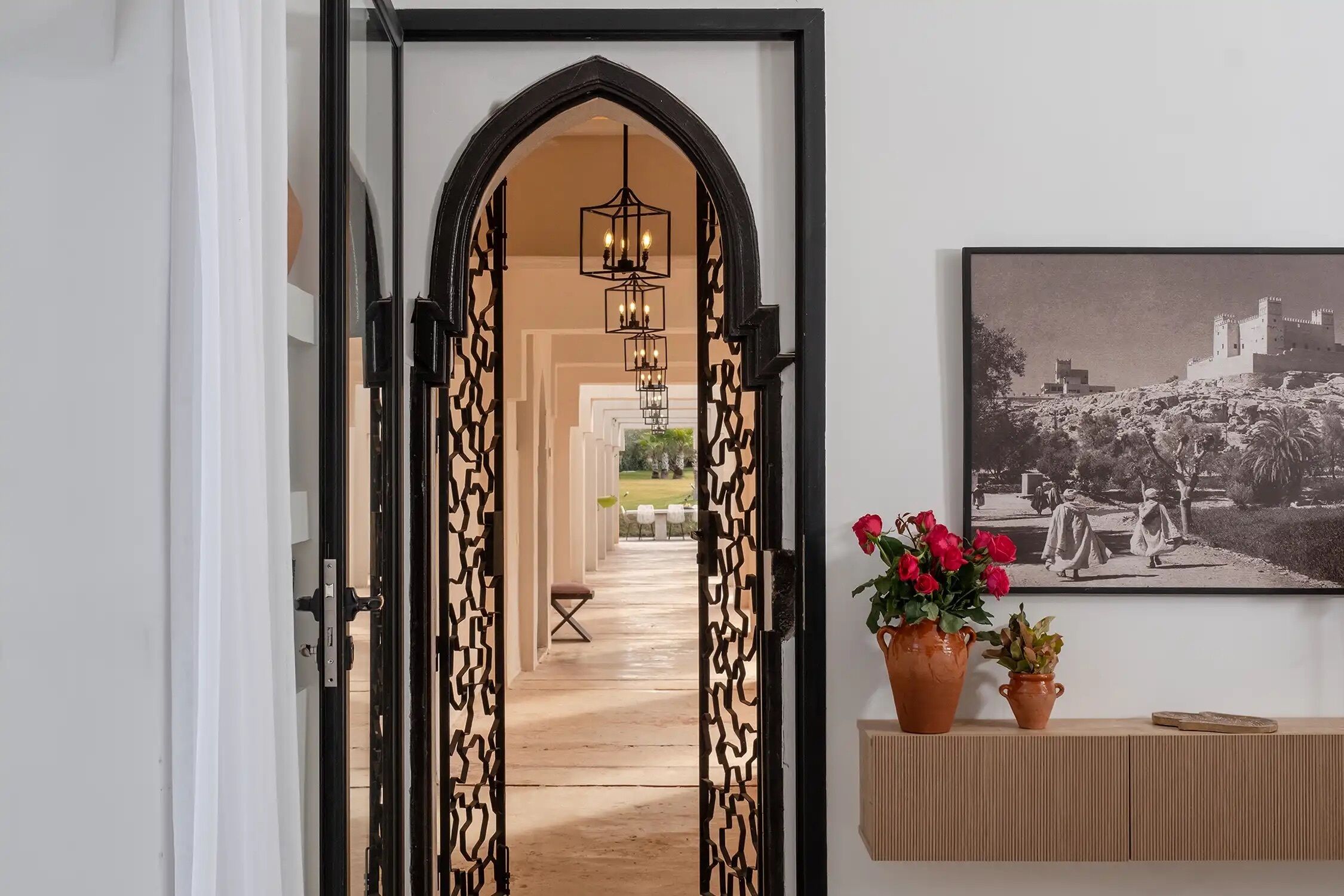 A hallway with an arched doorway, a hanging light fixture, a framed photograph on the wall, and two potted plants.
