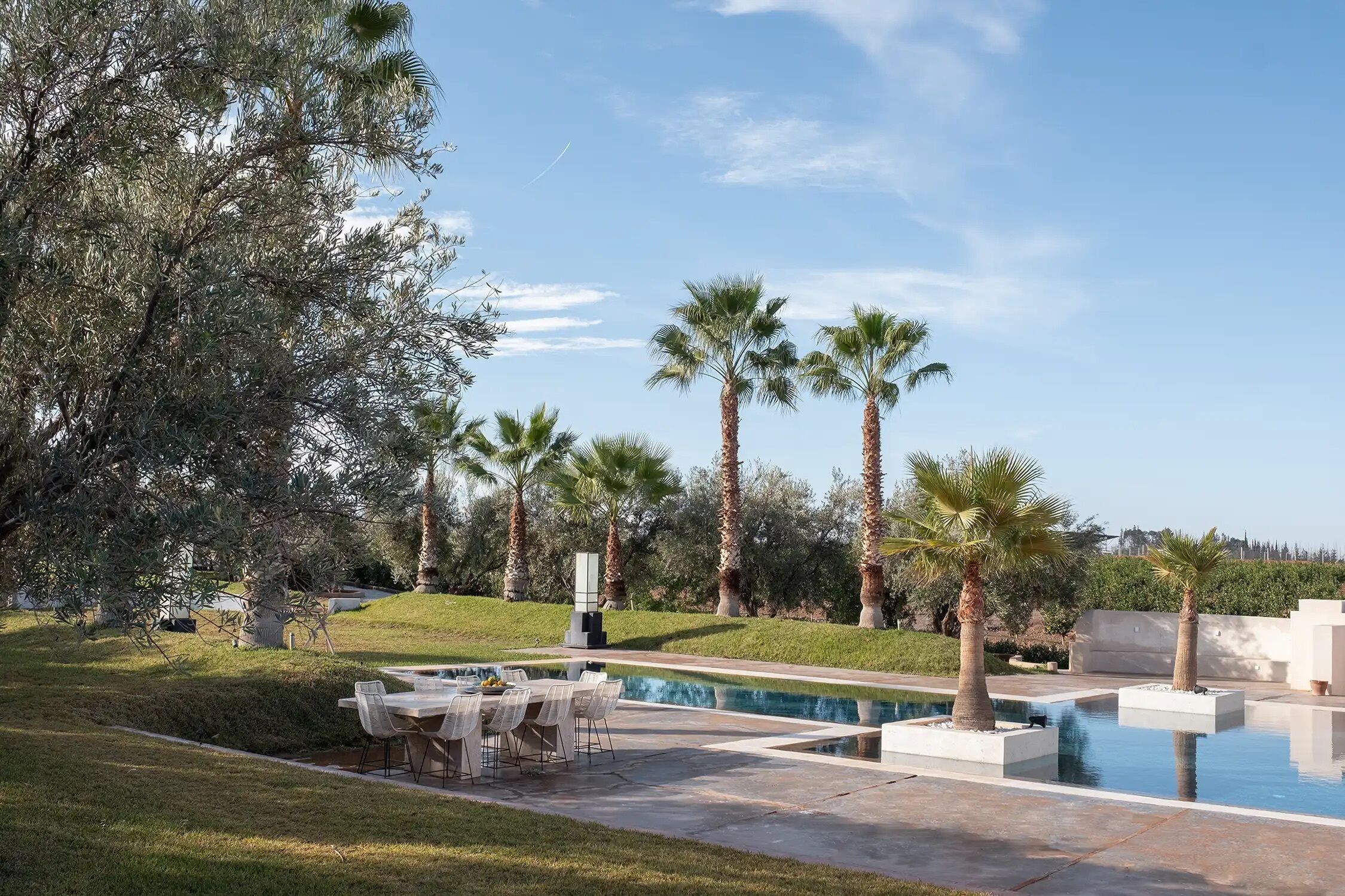 A pool area with a table and chairs, surrounded by palm trees and a grassy landscape.