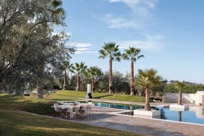 A pool area with a table and chairs, surrounded by palm trees and a grassy landscape.